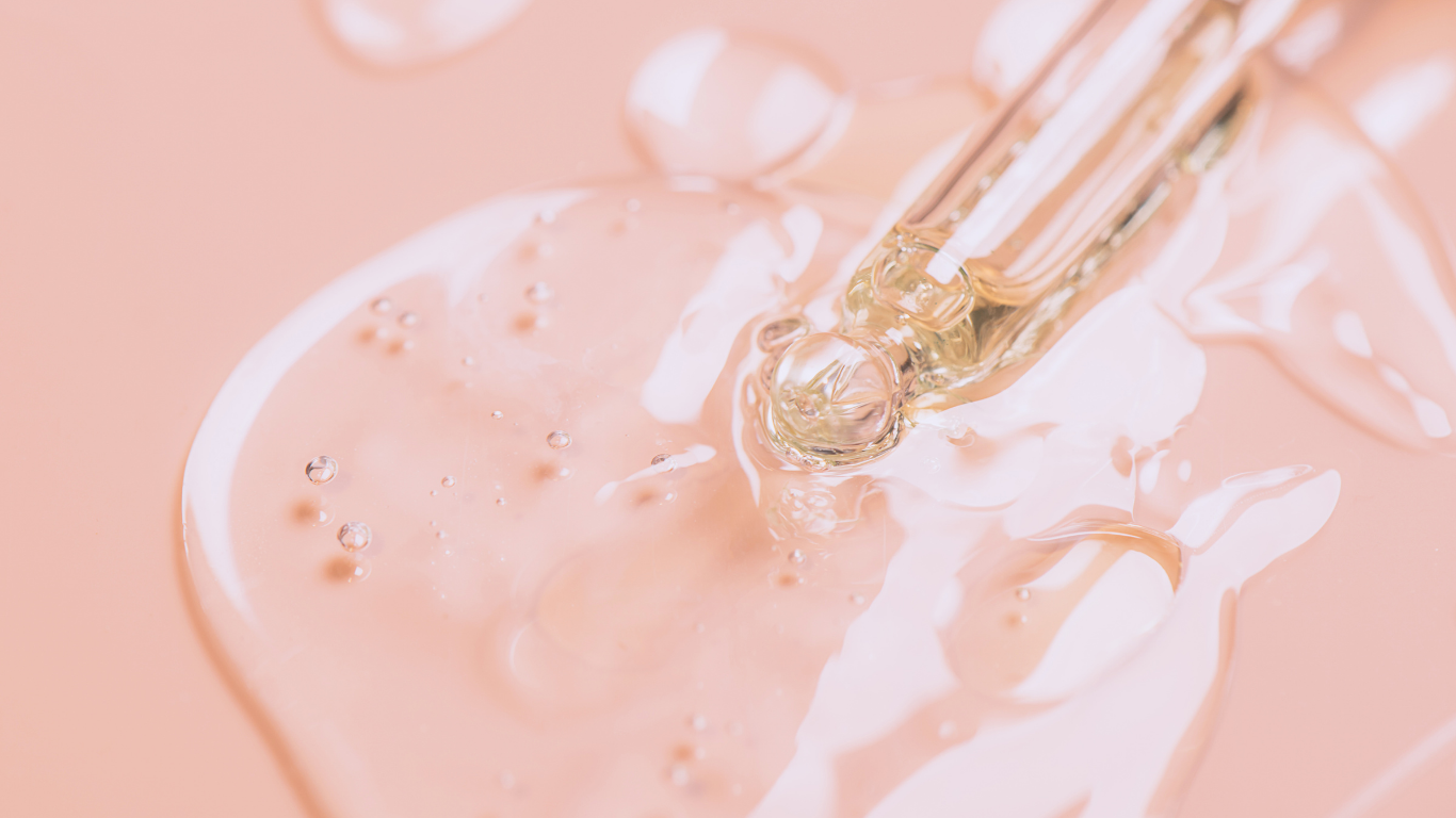 A close-up view of a clear liquid with small bubbles and a glass dropper, set against a soft pink background.