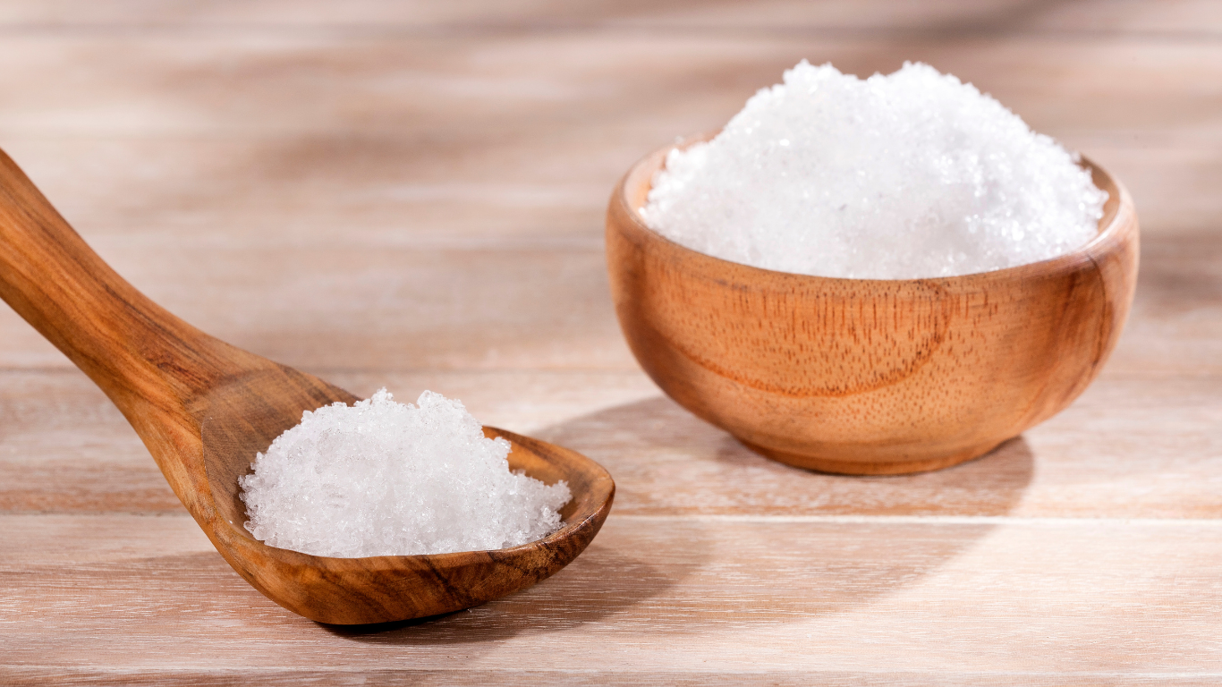 A wooden spoon and a wooden bowl filled with white crystalline flakes, magnesium chloride, placed on a light wooden surface.