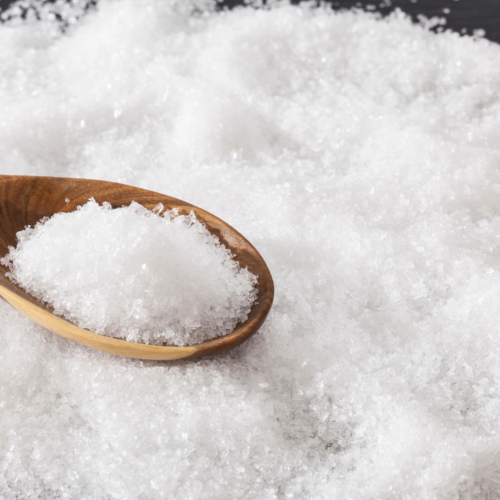 A wooden spoon filled with white crystalline flakes resting on a larger pile of similar crystals, magnesium chloride, spread across a dark surface.