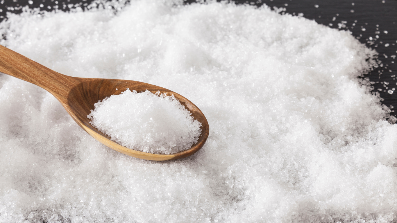 A wooden spoon filled with white crystalline flakes resting on a larger pile of similar crystals, magnesium chloride, spread across a dark surface.