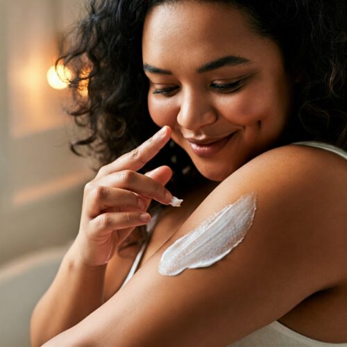 Person applying a thick layer of white body cream to the upper arm in a cozy indoor setting.