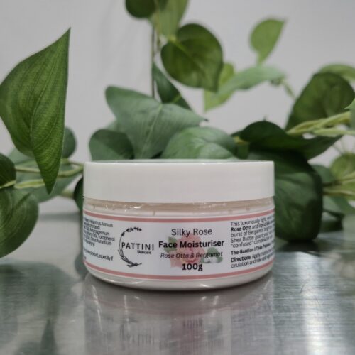 A Silky Rose Face Moisturiser 100g jar from Pattini Skincare placed on a reflective surface with green leaves in the background.