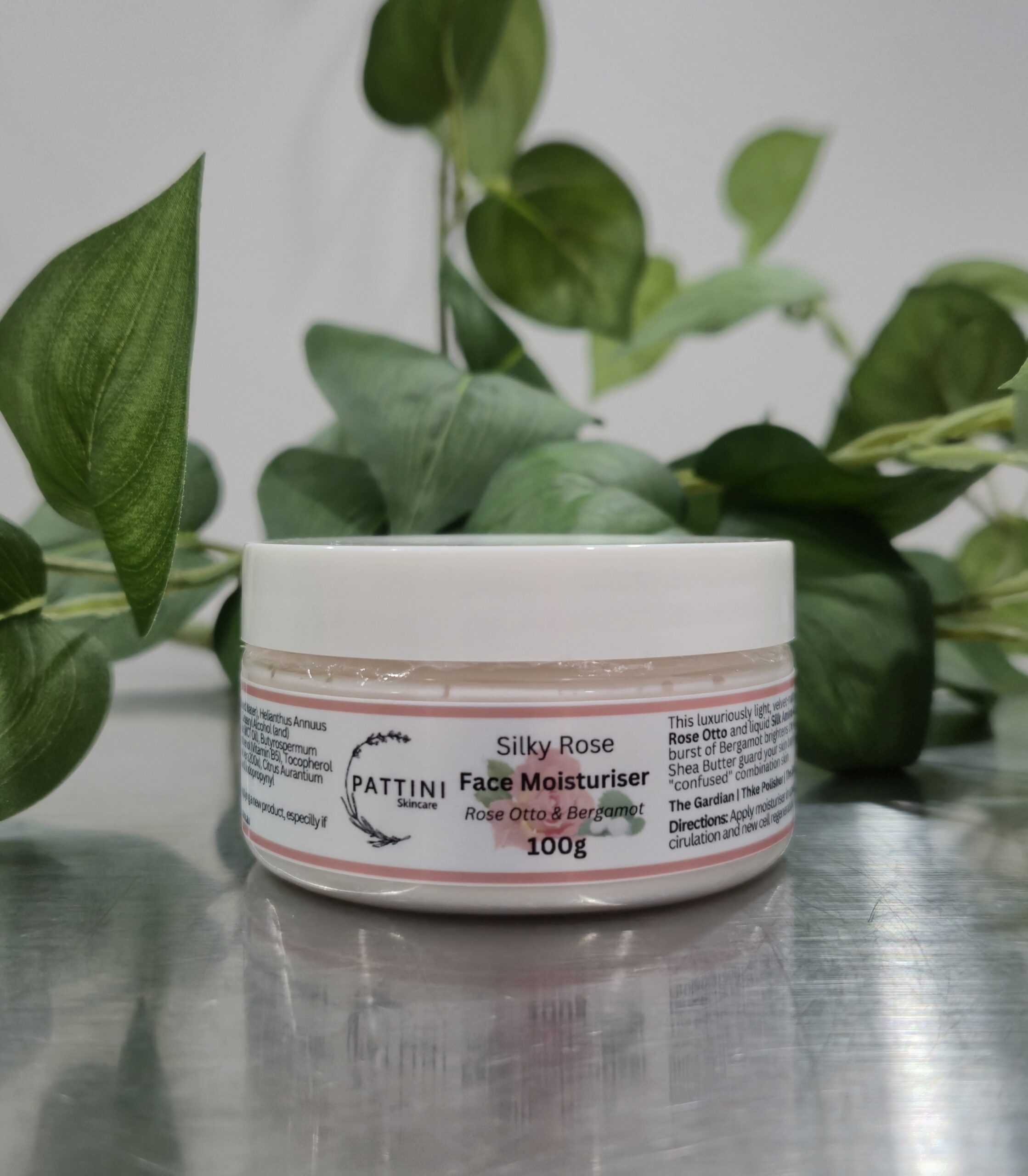 A Silky Rose Face Moisturiser 100g jar from Pattini Skincare placed on a reflective surface with green leaves in the background.